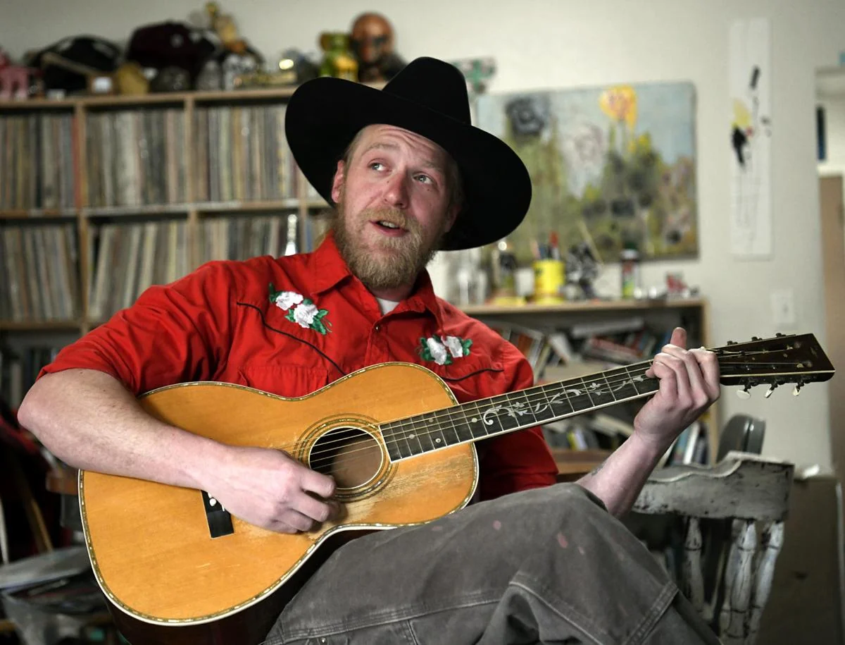 Aaron Jennings wearing a cowboy hat and playing an acoustic guitar.