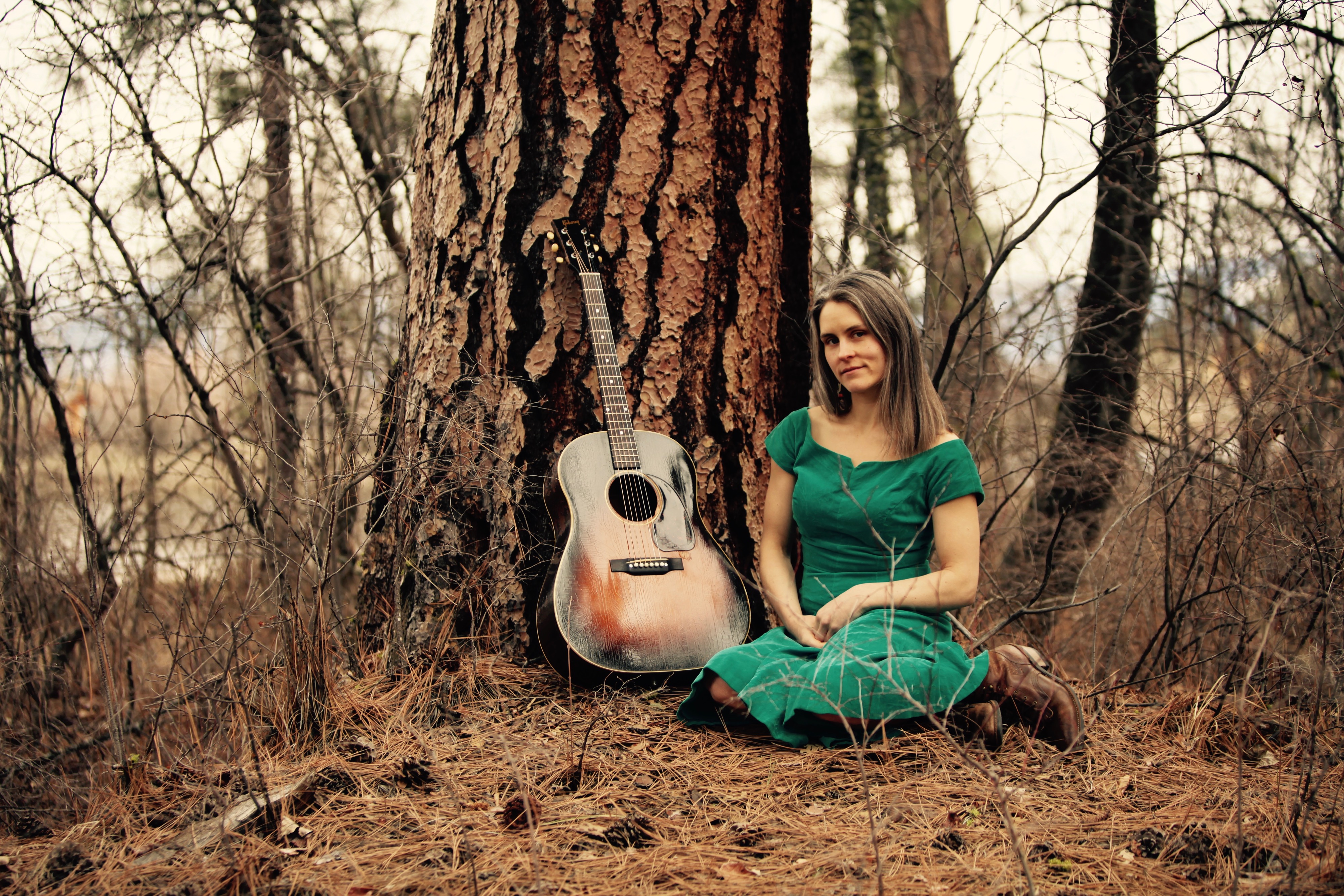 Britt Arnesen seated by a tree beside an acoustic guitar.